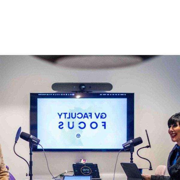 Two people smile while sitting on chairs. Microphones are sitting in front of them. A screen behind them says, "GV Faculty Focus."