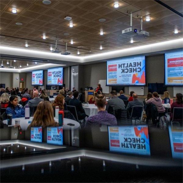 room of people seated at round tables, screens with Health Check around the room and reflected at bottom